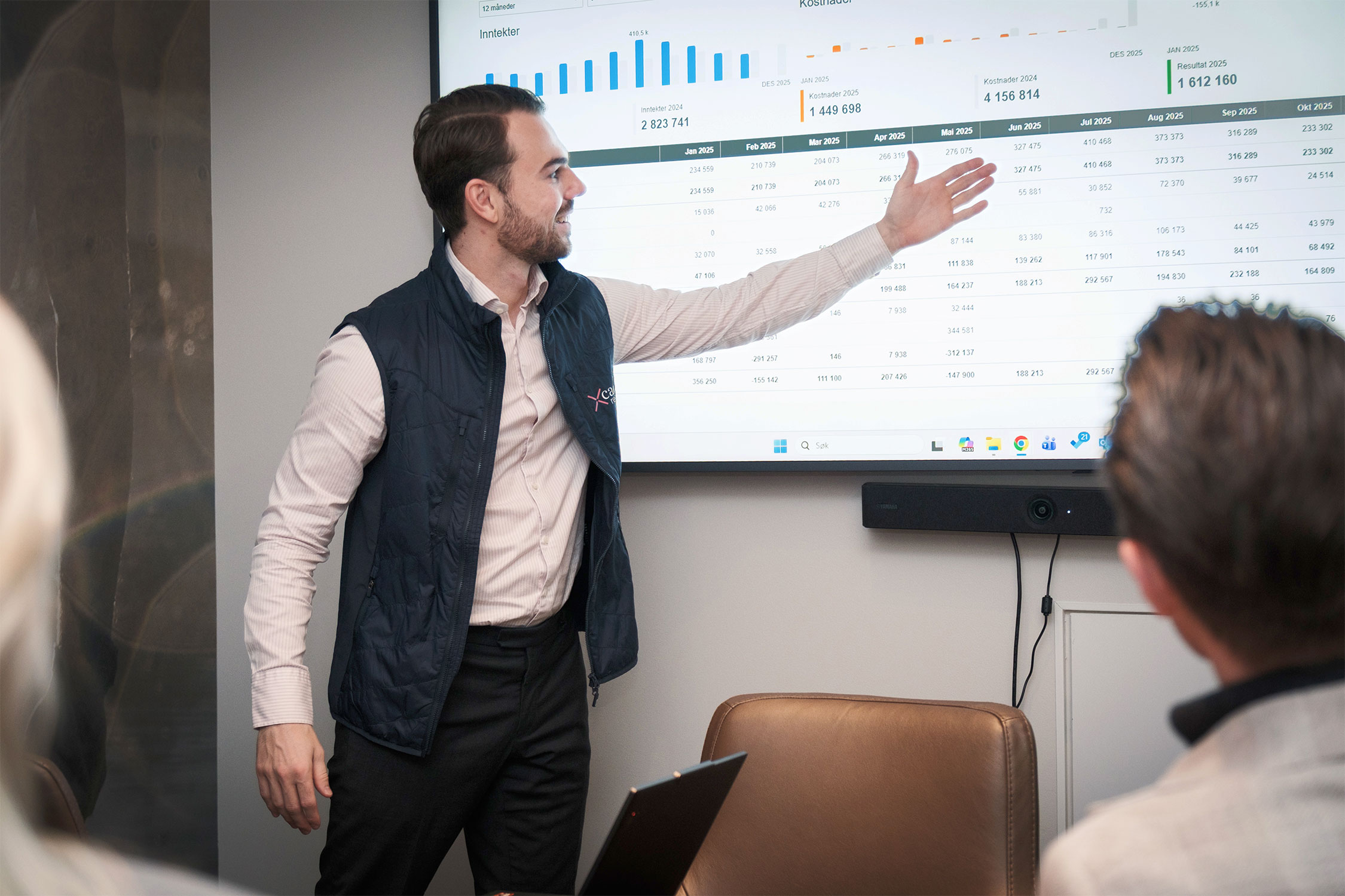 A person is presenting financial charts and data on a large screen in a meeting room, pointing at numbers on the display while others watch.