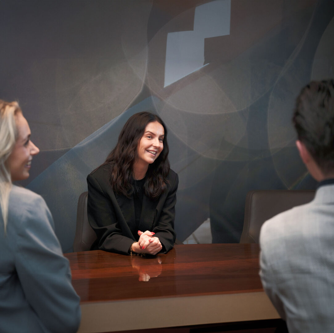 A woman is sitting at a conference table, smiling and speaking with two colleagues during a meeting in a modern office.