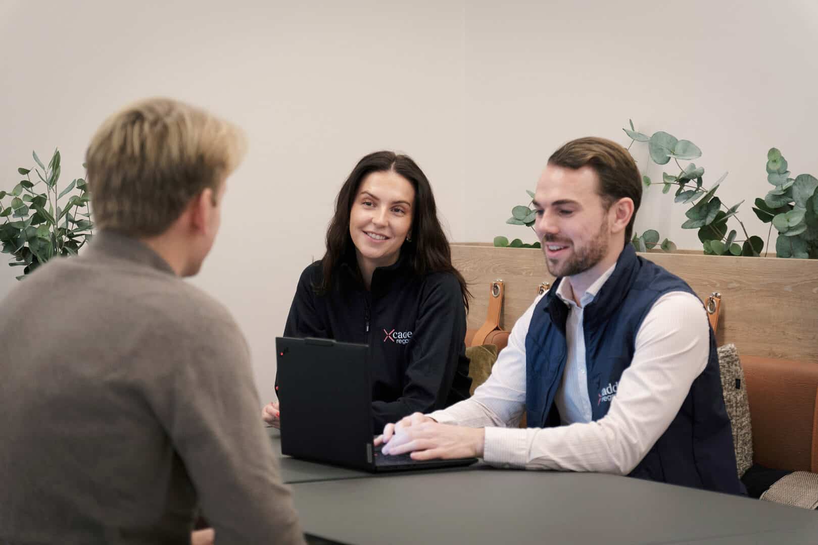 Three people are sitting at a table in an office, having a discussion. Two of them are using a laptop, and their clothing displays a logo of an accounting company. There are plants and wooden decor in the background.